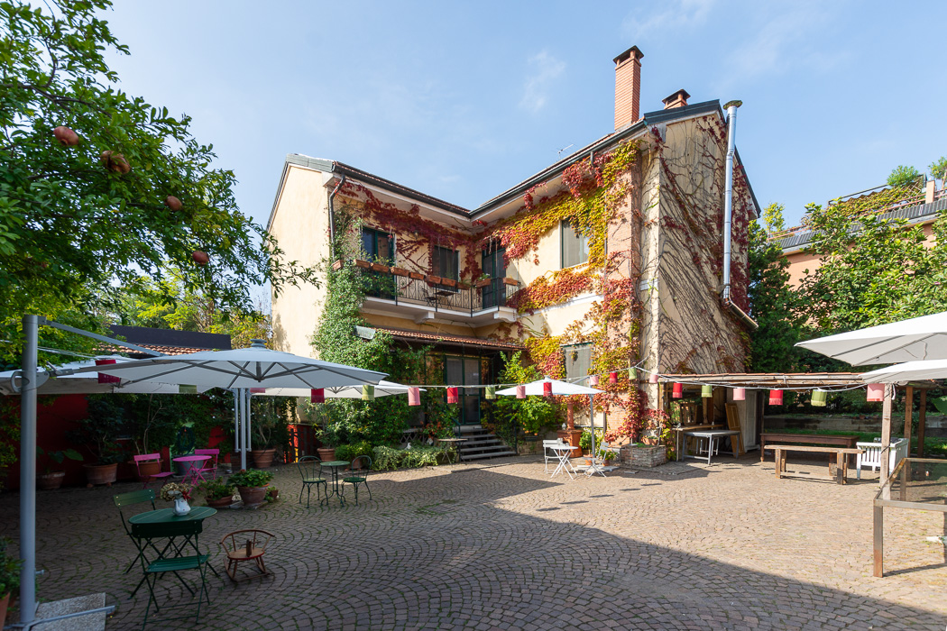 Il cortile della sede di Via Lucera 35 a Milano, con alberi, ombrelloni e l'edificio principale ricoperto di vite americana.